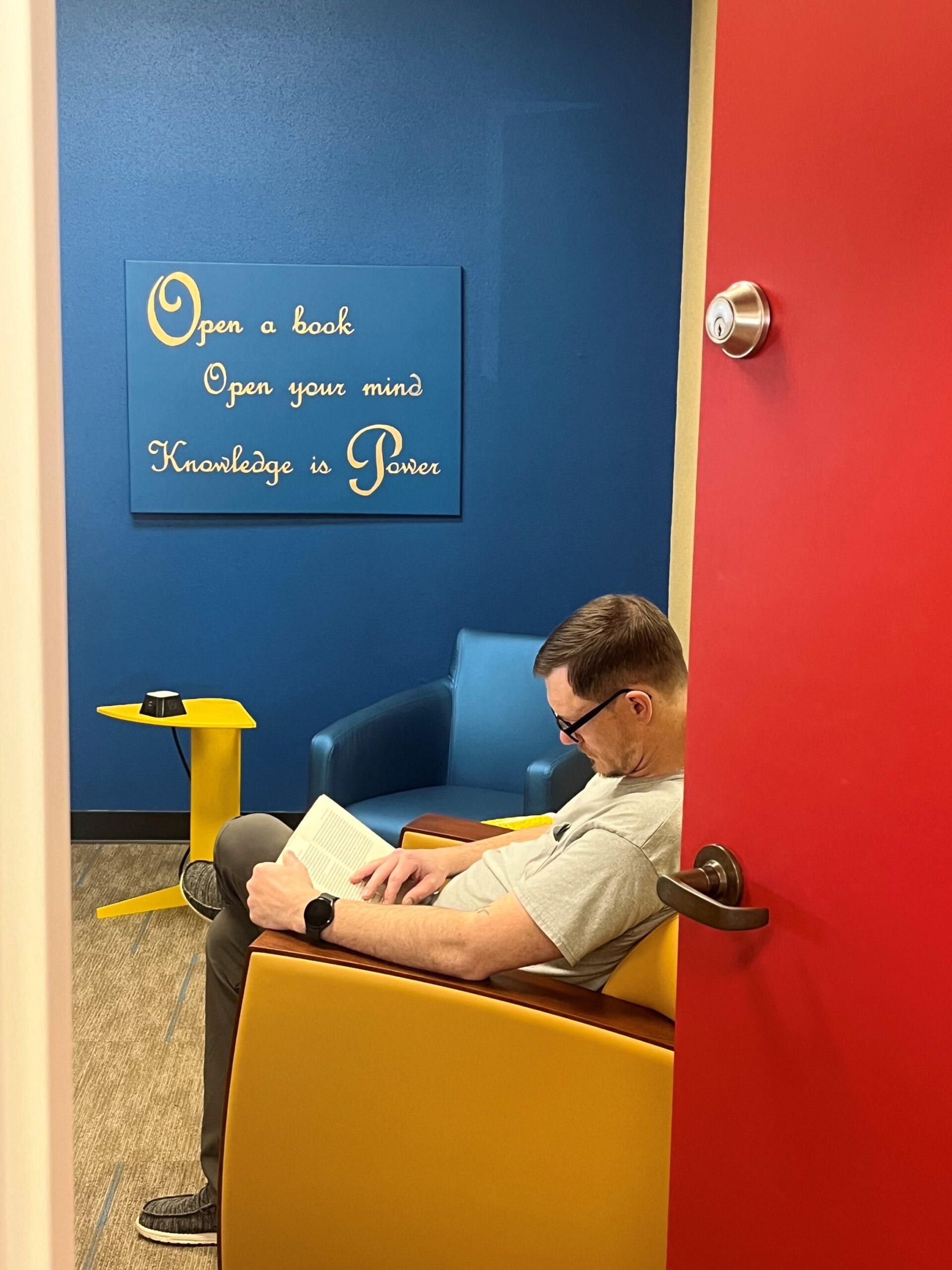 Man engrossed in a book in a bright blue library room featuring a poster that says: Open a book, open your mind, knowledge is power.